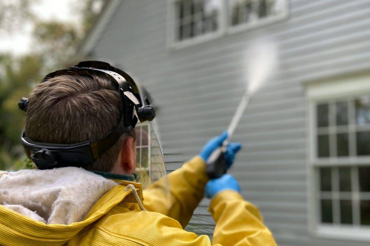 Person cleaning house siding with a pressure washer, wearing protective gear in a yellow raincoat.